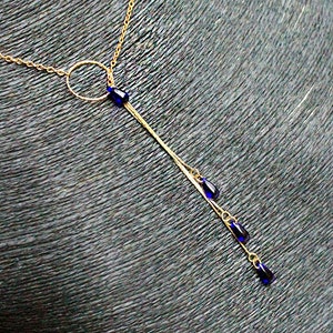 Op de afbeelding: Een gouden ketting met een cirkelvormige hanger en een lange, hangende ketting met vier blauwe druppelvormige kralen.