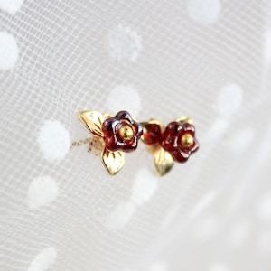 以下が含まれることがあります： 小さな金色の花のイヤリングのペア。各イヤリングは、金色の中心と2枚の金色の葉を持つ濃い赤色の花を特徴としています。イヤリングは柔らかい白い背景に配置されています。