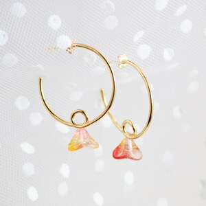 May include: Gold hoop earrings with a small, teardrop-shaped charm dangling from each hoop. The charms are made of clear glass with a red and yellow ombre effect.