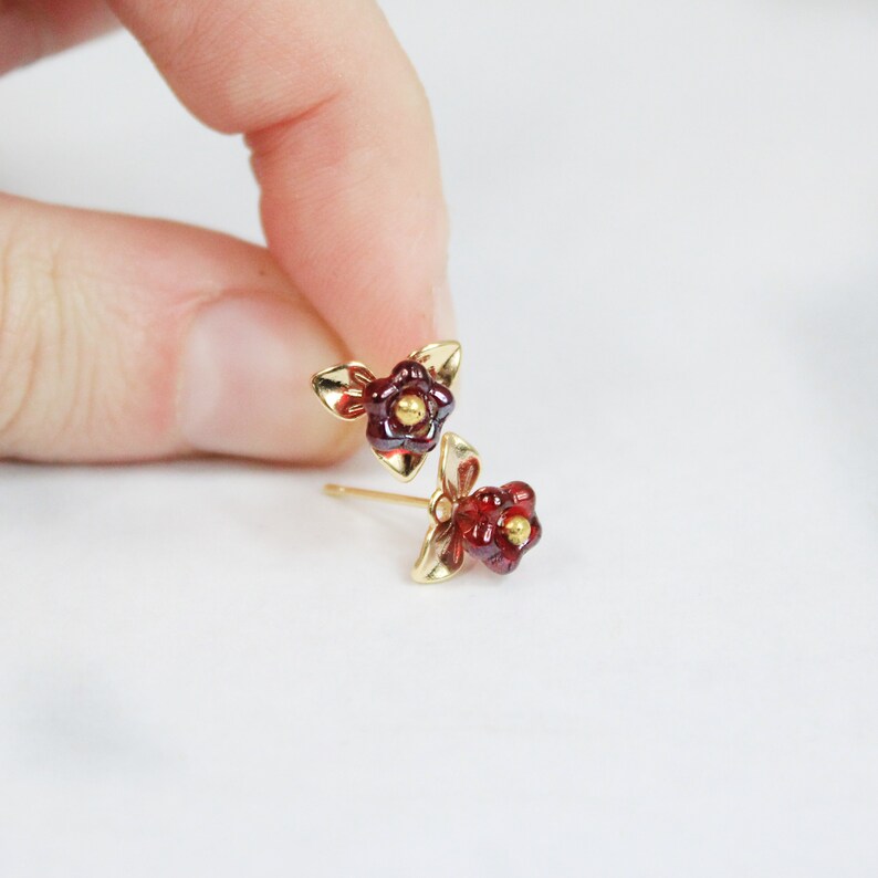 May include: A pair of gold-colored stud earrings, each featuring a small, red flower with a gold center and three gold leaves. The earrings are displayed against a white background.