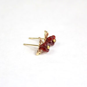 May include: A pair of gold-colored stud earrings with a flower design. The flower petals are a deep red color, and the center of the flower has a small gold-colored accent. The earrings are set against a white background.
