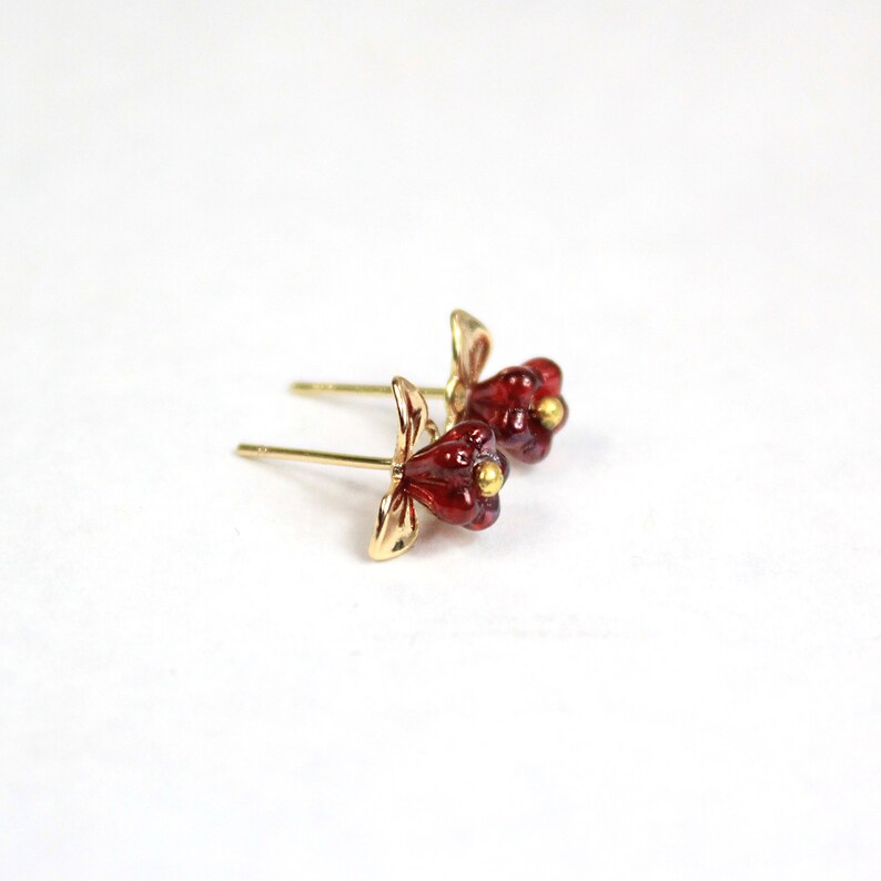 May include: A pair of gold-colored stud earrings with a flower design. The flower petals are a deep red color, and the center of the flower has a small gold-colored accent. The earrings are set against a white background.