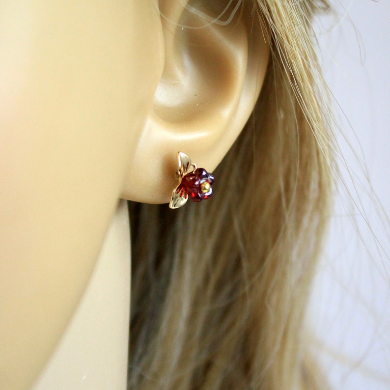 May include: A gold-colored floral stud earring with a red gemstone center and gold-colored leaves. The earring is small and delicate, designed to be worn on the ear. The gemstone has a faceted appearance.