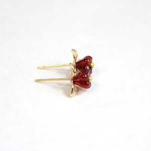 May include: A pair of gold-colored stud earrings featuring a flower design with red petals and a small gold-colored bead in the center. The earrings are set against a white background.