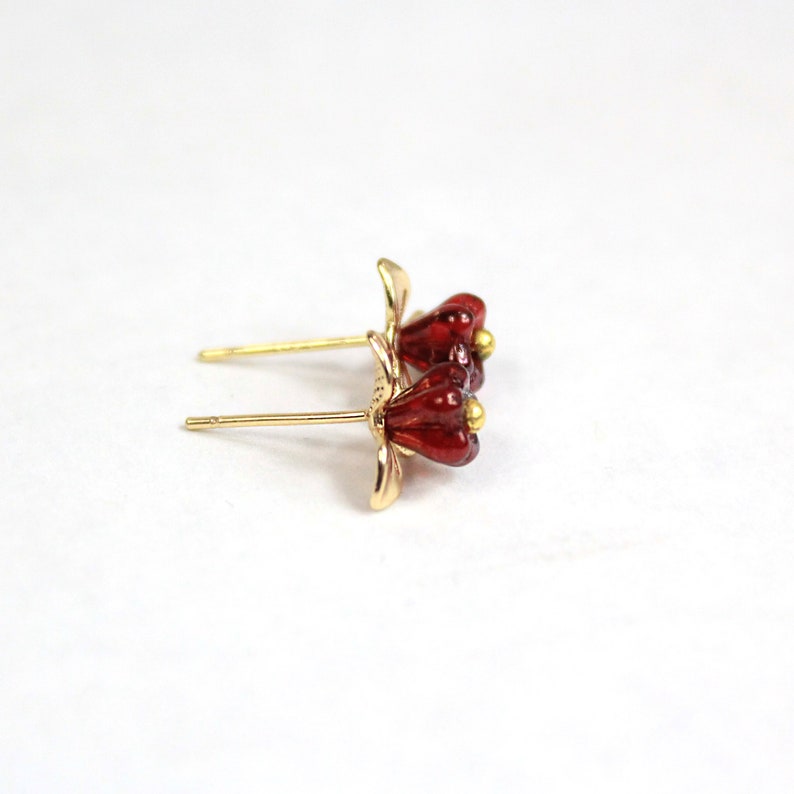 May include: A pair of gold-colored stud earrings featuring a flower design with red petals and a small gold-colored bead in the center. The earrings are set against a white background.