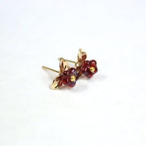 May include: A pair of gold-colored stud earrings featuring a floral design. Each earring has a red, flower-shaped center with a small, gold-colored accent in the middle. The earrings have a gold-colored leaf design.