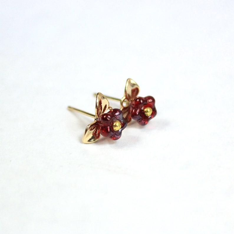 May include: A pair of gold-colored stud earrings featuring a floral design. Each earring has a red, flower-shaped center with a small, gold-colored accent in the middle. The earrings have a gold-colored leaf design.