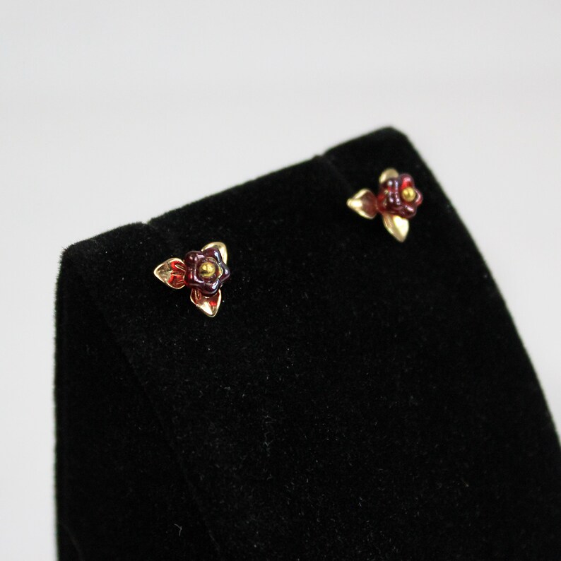 May include: A pair of gold-toned stud earrings, each featuring a small, stylized flower design. The flowers have burgundy petals and a golden center. The earrings are displayed on a black velvet surface against a white background.