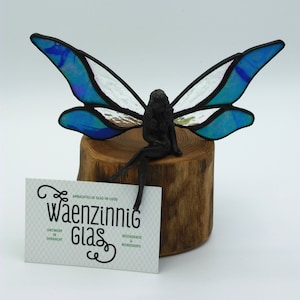 Może przedstawiać: Szklana wróżka z niebieskimi skrzydłami siedzi na drewnianym pniu. Wróżka jest wykonana z metalu i ma szczegółowy wzór. Skrzydła są tęczowe i mają niebiesko-fioletowy odcień. Drewniany pień jest naturalny i ma szorstką fakturę. Wróżka jest pięknym dziełem sztuki i byłaby wspaniałym dodatkiem do każdego domu.