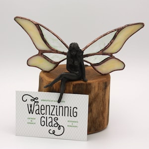 Może przedstawiać: Szklana wróżka z żółtymi i zielonymi skrzydłami siedzi na drewnianej podstawie. Wróżka jest czarna i ma delikatną, kobiecą formę. Podstawa jest w naturalnym kolorze drewna i ma szorstką fakturę. Wróżka jest kapryśnym i ozdobnym dziełem sztuki.