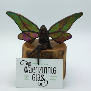 Może przedstawiać: Szklana wróżka z tęczowymi zielonymi i fioletowymi skrzydłami siedzi na drewnianym pniu. Wróżka trzyma wizytówkę z napisem "Waenzinnig Glas" z tekstem "Ontwerp in Opdracht" i "Restauratie & Workshops".