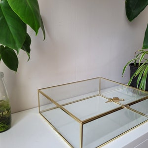 Może przedstawiać: Przezroczysta szklana gablotka z pozłacanym metalowym obramowaniem. Gablotka jest pusta i zawiera mały klucz w środku. Gablotka stoi na białej powierzchni.