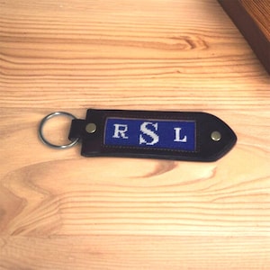 May include: A dark brown leather keychain with a blue embroidered panel displaying the initials "RSL" in white. The keychain has a silver metal ring and is secured with two small rivets.