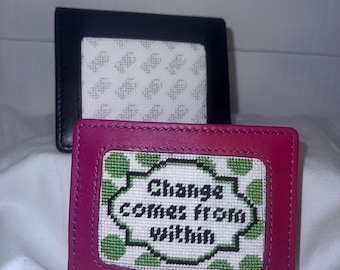 May include: Two leather wallets, one black and one magenta. The magenta wallet features a stitched design with the words "Change comes from within" in a white and green pattern. The black wallet is in the background.