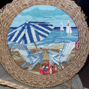 Puede incluir: Bolso redondo de paja tejida con una escena de playa bordada a punto de cruz. La escena representa una sombrilla a rayas azules y blancas, sillas de playa, veleros y una cesta de picnic en una playa de arena. El bolso tiene una correa tejida.