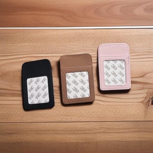 May include: Three rectangular card holders in black, brown, and pink. Each has a pocket on the front with a 3M adhesive backing. The card holders are displayed on a wooden surface.