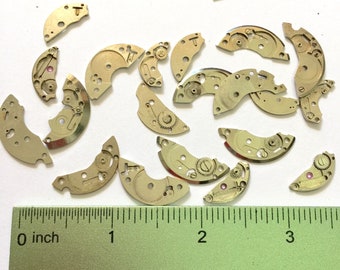 Könnte beinhalten: Eine Sammlung von 20 goldfarbenen Metall-Uhrenteilen, einige mit Zahnrädern, auf einer weißen Oberfläche mit einem grünen Lineal in Zoll.