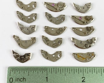 May include: A collection of small, silver-colored watch movement parts, including plates and gears, arranged on a white surface. A green ruler with inch measurements is at the bottom of the image.