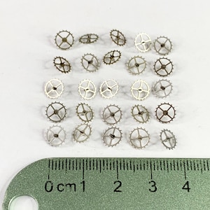 May include: A selection of small, metallic watch gears of various sizes and designs, scattered on a white surface. A green ruler with centimetre markings is in the foreground, providing scale. The gears have a spoked wheel design with small teeth.
