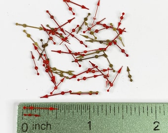 Puede incluir: Primer plano de un montón de manecillas de reloj rojas y doradas, que miden aproximadamente 2,5 cm de largo. Las manecillas están esparcidas sobre una superficie blanca con una regla para la escala.