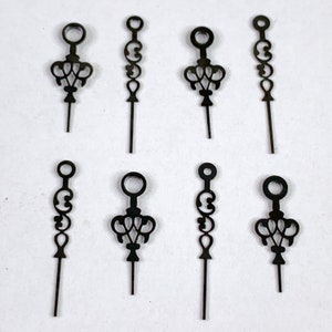 Puede incluir: Ocho agujas de reloj de metal negro con diseños ornamentados. Las agujas son todas del mismo tamaño y forma, con un pequeño bucle en la parte superior y un tallo largo y delgado.