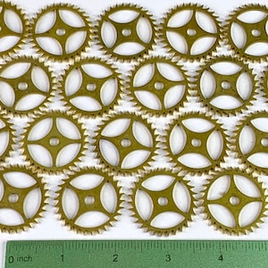 Peut inclure: Gros plan sur 16 engrenages en métal de couleur dorée avec un motif en étoile à quatre branches. Les engrenages sont disposés en rangées sur une surface blanche. Une règle est visible en bas de l'image, avec des graduations en pouces.