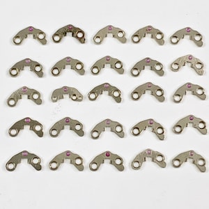 May include: Twenty-five silver-toned metal pieces with a small pink dot in the center of each piece. The pieces are shaped like a crescent moon with two small holes on each side.