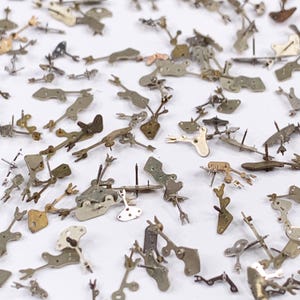 May include: A collection of small, vintage watch parts scattered across a white surface. The parts are made of metal and have a variety of shapes, including levers, gears, and other intricate components. The parts are in shades of silver, gold, and bronze.