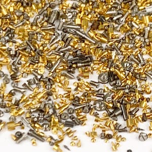 May include: A close-up of a collection of small metal components, including screws, bolts, and pins. The hardware is in gold and silver tones. The items are scattered, suggesting a collection of watchmaking or jewellery-making supplies.