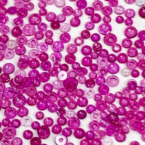 May include: A collection of small, round, translucent beads in varying shades of pink and magenta. The beads are scattered across a white surface, creating a textured, colorful pattern. The sizes of the beads vary, adding visual interest.