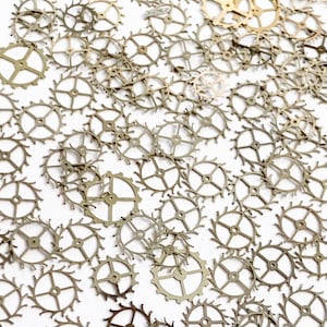 May include: A large quantity of small, gold-colored metal gears with intricate designs. The gears are scattered on a white surface.