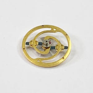 May include: A gold-colored watch movement with a spiral spring and a small red dot.