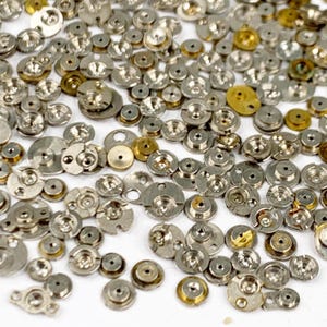 May include: A collection of small, silver-toned watch parts, including various sizes of circular and disc-shaped components. Some pieces have a gold hue, and all are scattered across a white surface. The parts have a mechanical appearance.