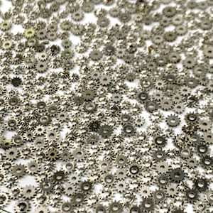 Puede incluir: Una gran cantidad de pequeños engranajes metálicos de color plateado esparcidos sobre una superficie blanca. Los engranajes son todos del mismo tamaño y forma, y tienen un acabado ligeramente texturizado.