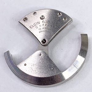 May include: A silver-toned watch movement with the text "ELGIN 618 U.S.A. S 6 7 7 2 9 9 18 JEWELS ADJUSTED" on the face.