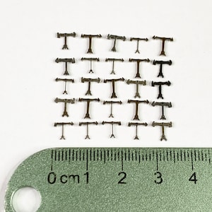 Op de afbeelding: Vijfentwintig kleine metalen stukjes in de vorm van een T met twee pijlen die in tegengestelde richtingen wijzen. De stukjes zijn gerangschikt in vijf rijen van vijf. Een liniaal bevindt zich onderaan de afbeelding en laat zien dat de stukjes ongeveer 1 cm lang zijn.