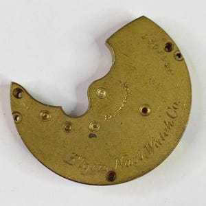 Może przedstawiać: Zabytkowa mosiężna płyta mechanizmu zegarka firmy Elgin Natl Watch Co. Zakrzywiona płyta ma kilka otworów i jest wygrawerowana nazwą firmy i numerem seryjnym. Płyta ma złoty kolor.