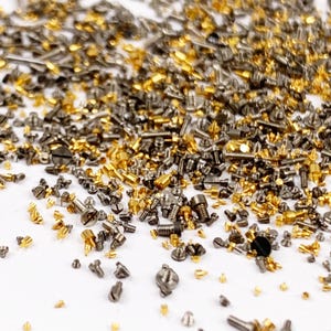 May include: A large collection of tiny metal screws and fasteners in gold and gunmetal grey tones. The assortment includes various head shapes, including flat, round, and slotted designs, scattered across a plain white surface.