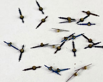 May include: A collection of 20 small, blue and black watch hands with gold accents. The hands are scattered on a white surface.