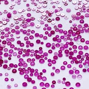 Op de afbeelding: Een verzameling van talloze kleine, ronde, magentakleurige lovertjes verspreid over een effen wit oppervlak. De lovertjes variëren in grootte en zijn voorzien van een centraal gat, met een glanzende en reflecterende afwerking.