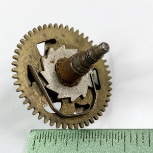 Pode incluir: Um conjunto de engrenagens de relógio vintage com uma engrenagem de latão, um centro prateado e um eixo de aço. A engrenagem tem muitos dentes e está ligeiramente manchada. Uma régua verde está em primeiro plano.