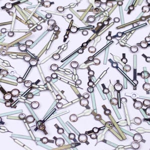 Puede incluir: Una colección de agujas de reloj en varios diseños y colores. Las agujas son plateadas, negras y blancas, algunas con un brillo verde o azul luminoso. El surtido incluye diferentes estilos para horas, minutos y segundos.