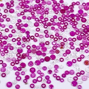 May include: A collection of small, clear, and magenta glass beads and settings. The beads are in various shapes, including round and ring-shaped, and are scattered across a white surface. The magenta beads vary in shade, creating a visually interesting pattern.