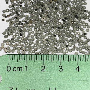 May include: A close-up of numerous tiny silver watch gears, scattered on a white background. The gears have a circular shape with toothed edges and a central hole. A green ruler with centimetre markings is at the bottom of the image.