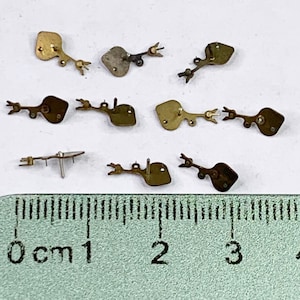 Puede incluir: Nueve piezas de reloj de metal dorado antiguo con diseños intrincados. Las piezas están dispuestas sobre una superficie blanca con una regla que mide en centímetros.