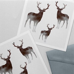 Puede incluir: Dos tarjetas blancas con una silueta marr&oacute;n de un ciervo con un bosque en su interior. Los ciervos son de diferentes tama&ntilde;os. Las tarjetas est&aacute;n sobre una superficie gris con un sobre gris.
