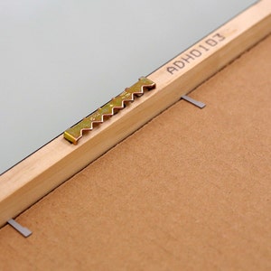 May include: Close-up of a wooden picture frame with a metal hanging hook and the text "ADHO103" printed on the back.