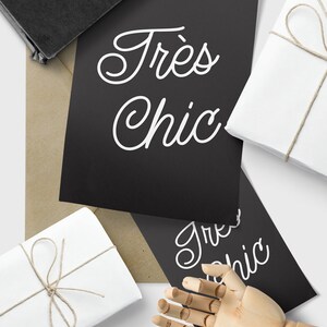 May include: Two black cards with white text that reads "Tr&egrave;s Chic" in a cursive font. The cards are on a brown paper surface with two white gift boxes wrapped with twine.