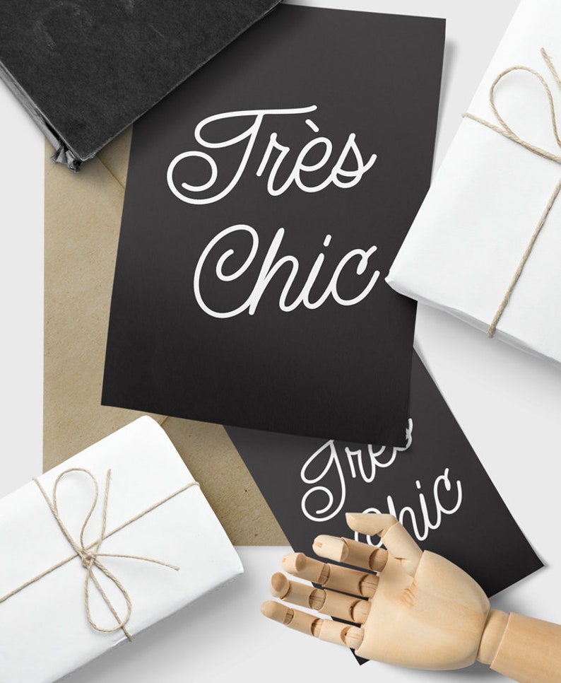 May include: Two black cards with white text that reads "Tr&egrave;s Chic" in a cursive font. The cards are on a brown paper surface with two white gift boxes wrapped with twine.