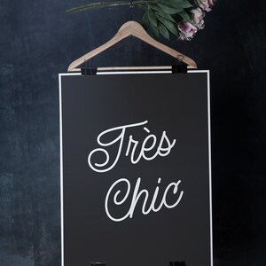 May include: A black and white poster with the text "Tr&egrave;s Chic" hanging on a wooden hanger against a dark blue background.